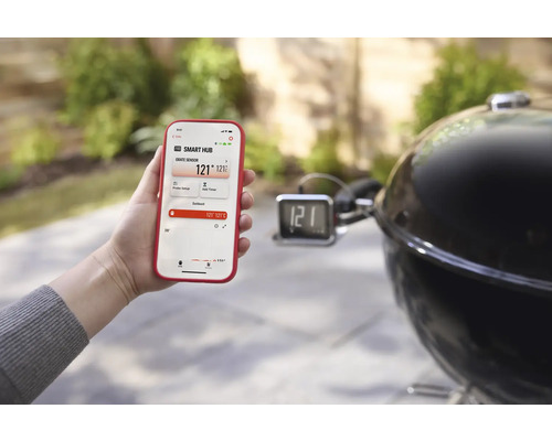 Eine Person hält ein Smartphone mit der Weber Connect App neben einem Holzkohlegrill.