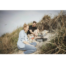 Ein Paar grillt mit einem Elektrogrill in den Dünen, umgeben von Strandhafer und mit Getränken und Kissen.