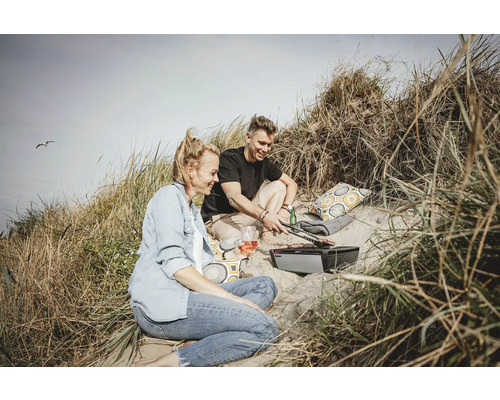 Ein Paar grillt mit einem Elektrogrill in den Dünen, umgeben von Strandhafer und mit Getränken und Kissen.