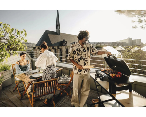 Personen grillen auf einer Dachterrasse mit Gasgrill und Holzzange, während im Hintergrund eine Kirche zu sehen ist.