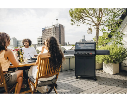 Personen sitzen auf einer Dachterrasse am Tisch und grillen mit einem Gasgrill.