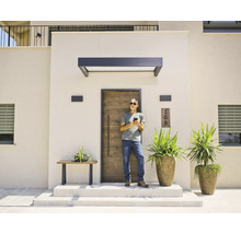 Moderner Eingangsbereich mit Haustür in Holzoptik, dunklem Vordach und Außenwandleuchten. Ein Mann steht vor der Tür und nutzt sein Smartphone.