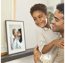 Ein Vater hält seinen lächelnden Sohn im Arm, beide betrachten einen auf einem Regal stehenden digitalen Bilderrahmen mit einem Familienfoto.