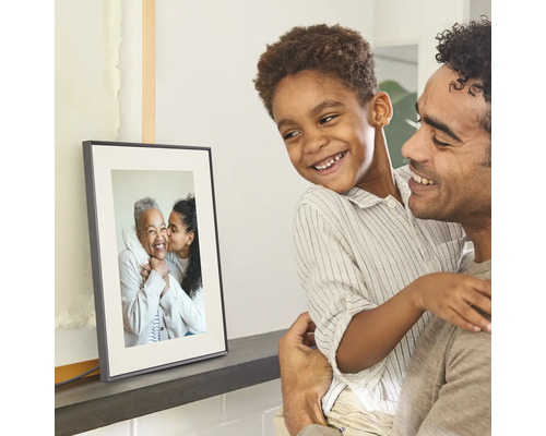 Ein Vater hält seinen lächelnden Sohn im Arm, beide betrachten einen auf einem Regal stehenden digitalen Bilderrahmen mit einem Familienfoto.
