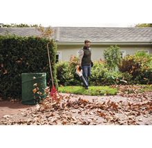 Ein Mann benutzt einen Laubbläser im Garten, neben einem Laubsack und einem Rechen.