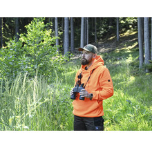 Mann mit Fernglas, orangefarbenem Hoodie und Tarnkappe im Wald.