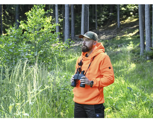 Mann mit Fernglas, orangefarbenem Hoodie und Tarnkappe im Wald.
