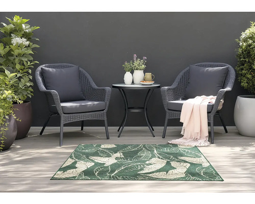 Gartenszene mit zwei Rattansesseln, kleinem Tisch und einem grünen Teppich mit Blattmuster