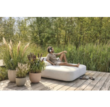 Mann entspannt auf einer Gartenliege auf einer Holzterrasse, umgeben von Gräsern und Pflanzen in Pflanzgefäßen.