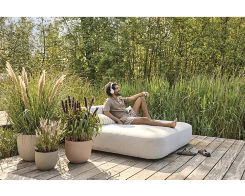 Mann entspannt auf einer Gartenliege auf einer Holzterrasse, umgeben von Gräsern und Pflanzen in Pflanzgefäßen.