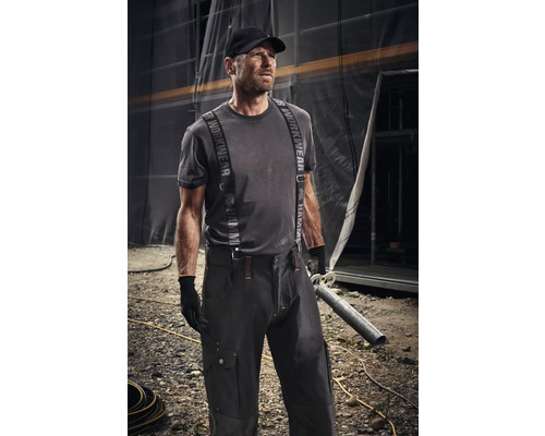 Mann in Arbeitskleidung mit Hosenträgern und Mütze hält ein Rohr