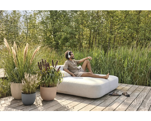 Szene im Garten mit einem Mann, der auf einem Lounge-Sofa liegt, umgeben von Ziergräsern in Pflanzgefäßen.