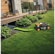 Ein Rasenmähroboter fährt auf einer Grünfläche zu seiner Ladestation unter einer Holzterrasse.