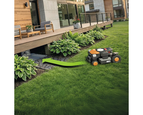 Ein Rasenmähroboter fährt auf einer Grünfläche zu seiner Ladestation unter einer Holzterrasse.