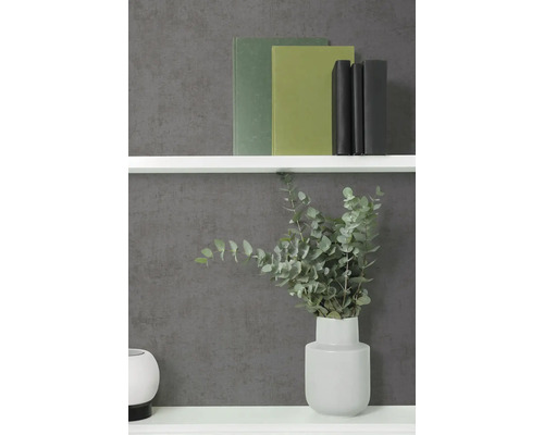 Vliestapete in Anthrazit mit Betonoptik und feiner Struktur, kombiniert mit einem weißen Regal, Büchern und einer Vase mit Eukalyptus.
