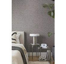 Graue Vliestapete in Betonoptik in modernem Schlafzimmer mit Bett, silbernem Hocker, Lampe und großer Zimmerpflanze.