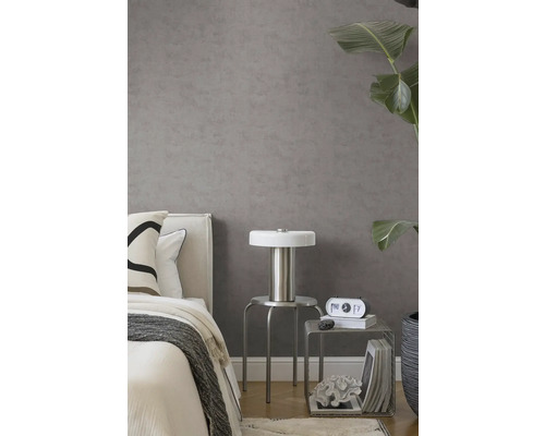 Graue Vliestapete in Betonoptik in modernem Schlafzimmer mit Bett, silbernem Hocker, Lampe und großer Zimmerpflanze.