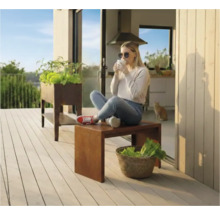 Szene auf einer Terrasse mit einem Hochbeet und einer Frau, die auf einem Hocker sitzt und ein Getränk genießt