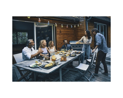 Gruppe von Personen isst und trinkt an einem Holztisch im Freien unter einer Lichterkette.