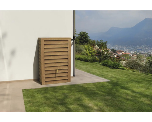 Wand-Regenspeicher in Holzoptik mit Zapfhahn an einer weißen Hauswand in einem hellen Garten mit Blick auf Berge.