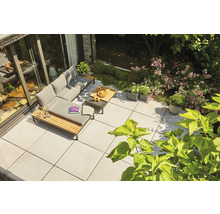 Moderne Terrasse mit hellgrauen Bodenplatten, L-förmiger Lounge-Gruppe mit grauen Polstern und Holzdetails inmitten eines Gartens.