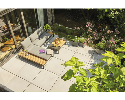 Moderne Terrasse mit hellgrauen Bodenplatten, L-förmiger Lounge-Gruppe mit grauen Polstern und Holzdetails inmitten eines Gartens.