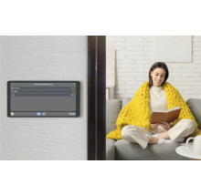 Smart Home Bedienpanel an der Wand mit Anzeige des Energieverbrauchs, im Hintergrund eine Frau auf einem Sofa in einem hellen Wohnzimmer.