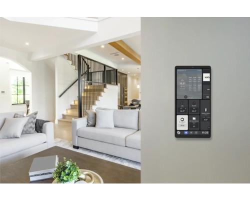 An der Wand montiertes Smart Home Bedienpanel in einem hellen, modernen Wohnzimmer mit Sofa und Treppe.