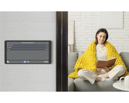 Smart Home Bedienpanel an der Wand mit Anzeige des Energieverbrauchs, im Hintergrund eine Frau auf einem Sofa in einem hellen Wohnzimmer.