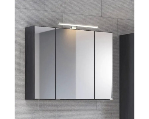 Dreitüriger Spiegelschrank fürs Badezimmer mit LED-Aufsatzleuchte und dunklem Korpus vor einer Wand in Steinoptik.