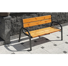 Gartenbank mit schwarzem Metallgestell und Sitz- sowie Rückenfläche aus Holzlatten auf einer gepflasterten Terrasse vor einer Steinmauer.