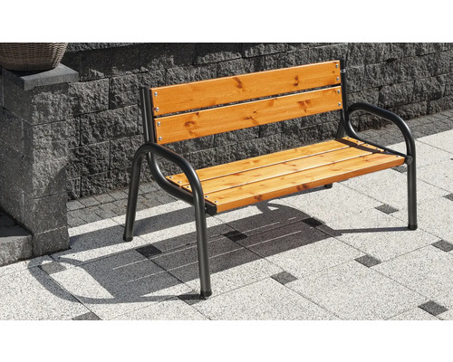 Gartenbank mit schwarzem Metallgestell und Sitz- sowie Rückenfläche aus Holzlatten auf einer gepflasterten Terrasse vor einer Steinmauer.