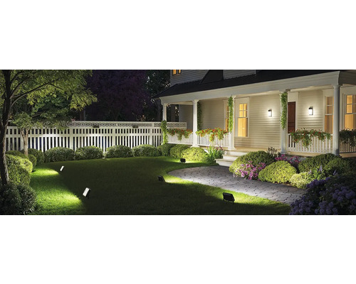 Beleuchteter Garten mit Rasen, Sträuchern, Zaun und Hausfassade mit Aussenleuchten.
