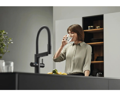Frau trinkt Wasser aus einem Glas neben einem Wasserhahn in einer Küche