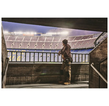 Ein Mann in Arbeitskleidung steht vor einem Stadion und blickt auf das Spielfeld.