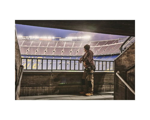 Ein Mann in Arbeitskleidung steht vor einem Stadion und blickt auf das Spielfeld.