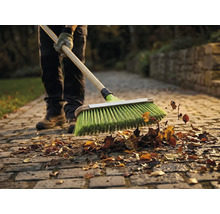 Eine Person kehrt Laub mit einem Besen auf einem Steinpflasterweg.