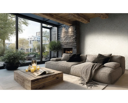 Helles Wohnzimmer mit grauem Stoffsofa, rustikalem Holztisch, Steinkamin und grossen Fenstern. Wand mit dezent gemusterter Tapete und Deckenbalken.