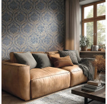 Helles Wohnzimmer mit blau-beiger Damasttapete hinter einem braunen Ledersofa mit Kissen und einem Holzfenster.