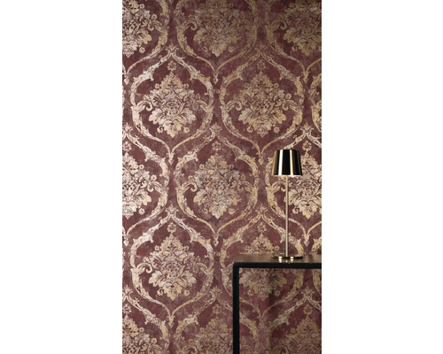 Vliestapete mit barockem Damast-Muster in Gold-Optik auf rotem Grund, dazu eine goldene Tischleuchte auf einem dunklen Tisch.