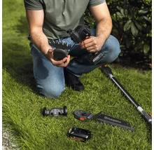 Eine Person hockt auf dem Rasen und hält eine Akku-Grasschere von Husqvarna, wobei verschiedene Aufsätze und ein Akku im Bild zu sehen sind.