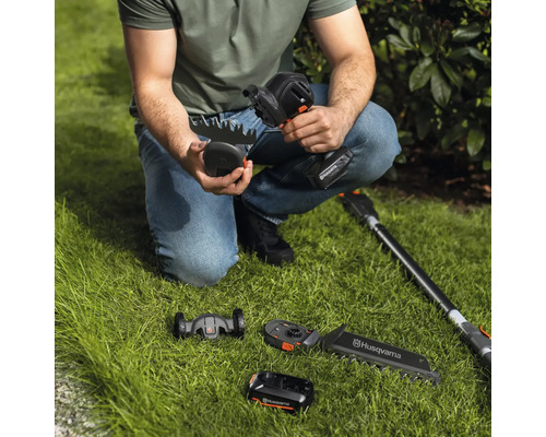 Eine Person hockt auf dem Rasen und hält eine Akku-Grasschere von Husqvarna, wobei verschiedene Aufsätze und ein Akku im Bild zu sehen sind.