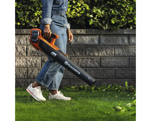 Eine Person trägt einen Akku-Laubbläser der Marke Husqvarna in einem Garten.