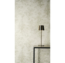 Vliestapete mit dezentem Baummuster und Waldmotiv in hellen Farbtönen hinter einer goldenen Tischleuchte auf einem schwarzen Tisch.