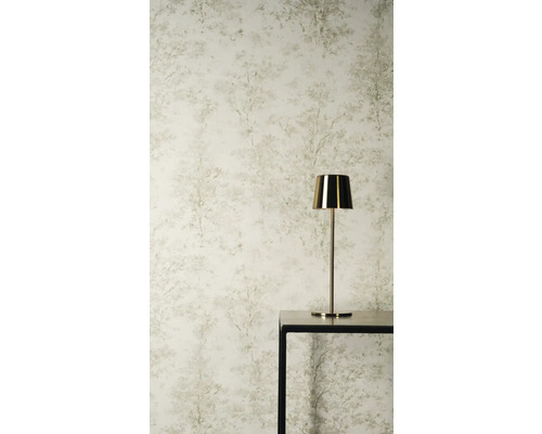 Vliestapete mit dezentem Baummuster und Waldmotiv in hellen Farbtönen hinter einer goldenen Tischleuchte auf einem schwarzen Tisch.