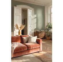 Helles Wohnzimmer mit salbeigrüner Wand, terrakottafarbenem Samtsofa, Holzkommode, Fischgrätparkett und einer großen Zimmerpflanze.