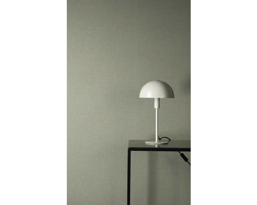 Strukturierte Vliestapete in Graugrün mit Leinenoptik, kombiniert mit einer weißen Tischleuchte auf einem schwarzen Metalltisch.