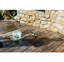 Reinigung einer Holzterrasse mit einer langstieligen Bürste und Osmo Holz-Entgrauer Kraft-Gel vor einer Steinmauer.