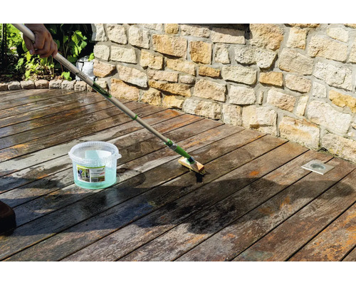 Reinigung einer Holzterrasse mit einer langstieligen Bürste und Osmo Holz-Entgrauer Kraft-Gel vor einer Steinmauer.