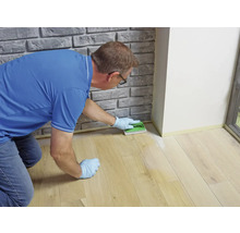 Eine Person kniet auf einem Holzfußboden neben einer Wand und trägt mit einem Applikator eine Bodenpflege auf.